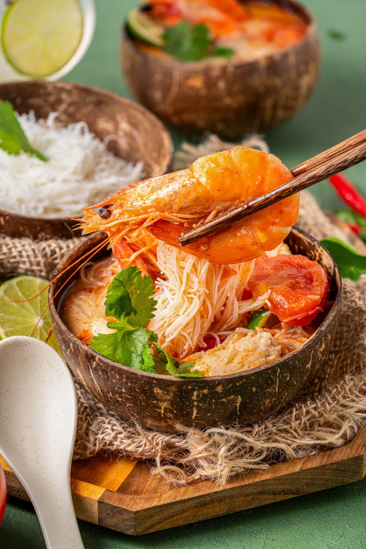 A pair of wooden chopsticks hold a large prawn over a bowl of vibrant red Tom Yum Soup.
