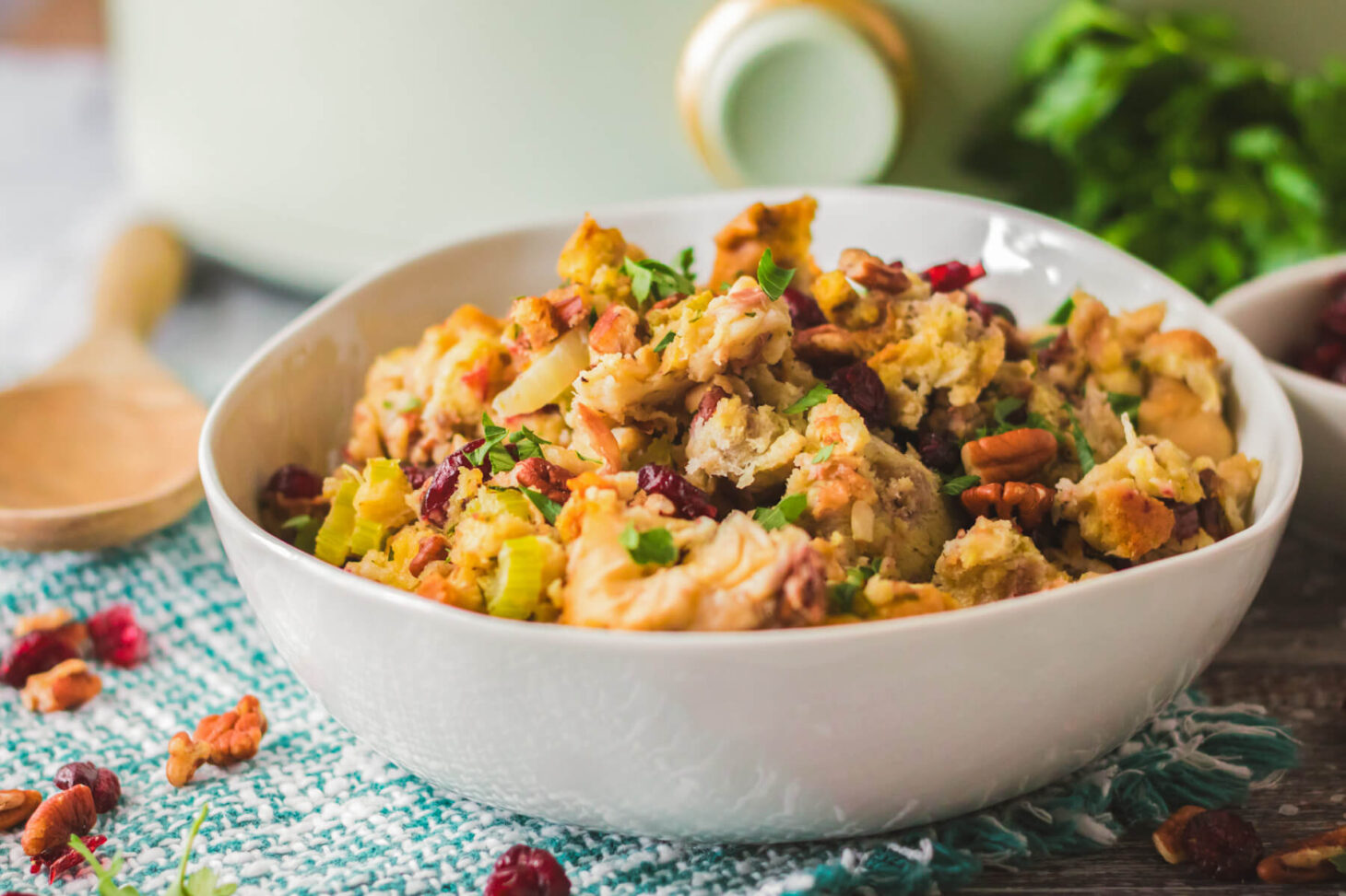 A white bowl with traditional bread dressing dotted with red dried cranberries, pecans, celery, and parsley in front of a slow cooker.