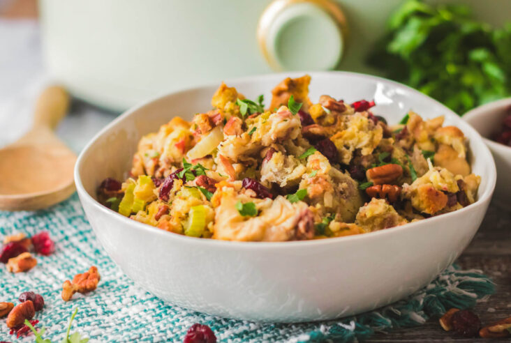 A white bowl with traditional bread dressing dotted with red dried cranberries, pecans, celery, and parsley in front of a slow cooker.