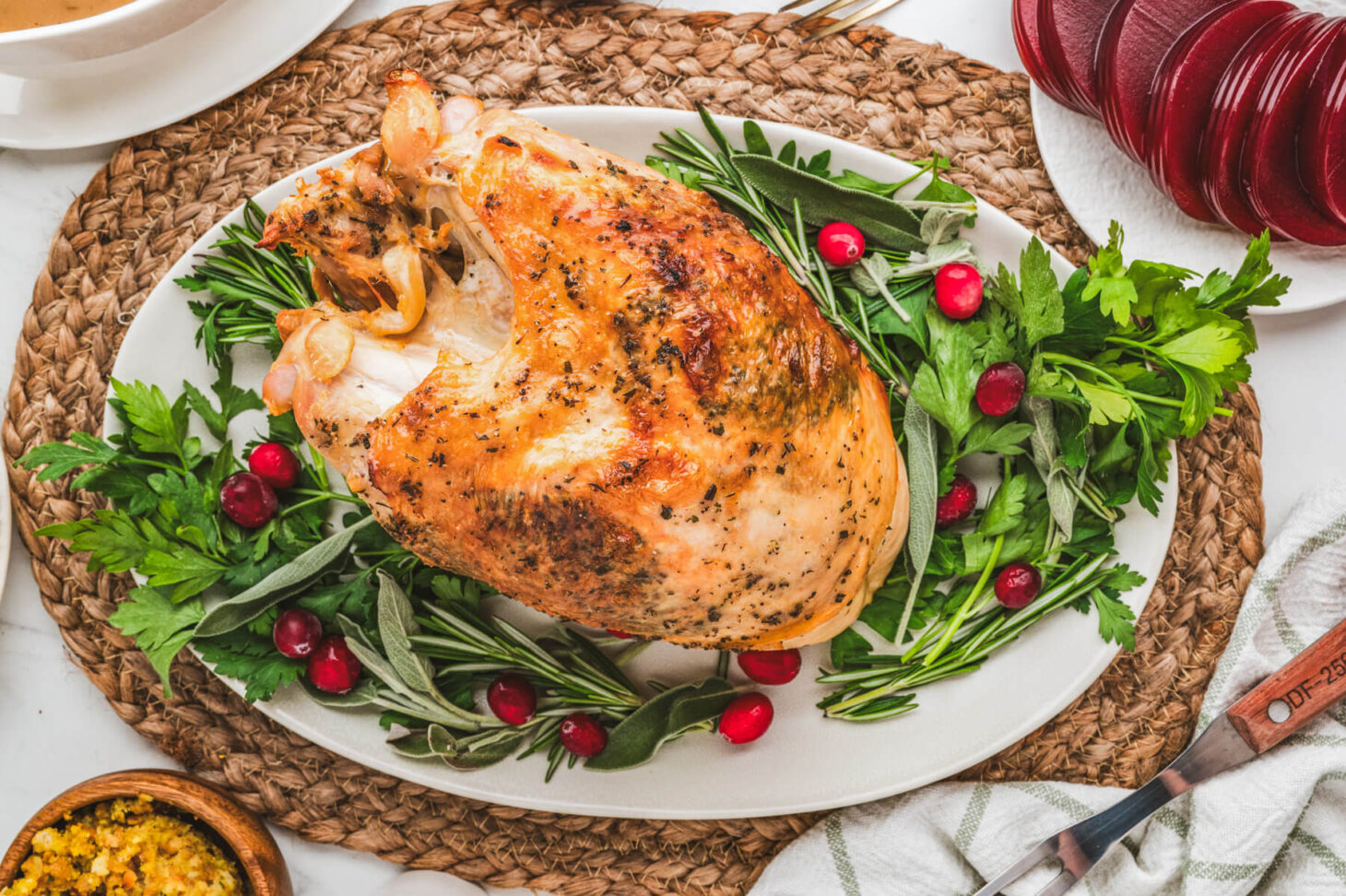 A golden brown crispy skin turkey breast on an oval platter surrounded by fresh herbs and cranberries.