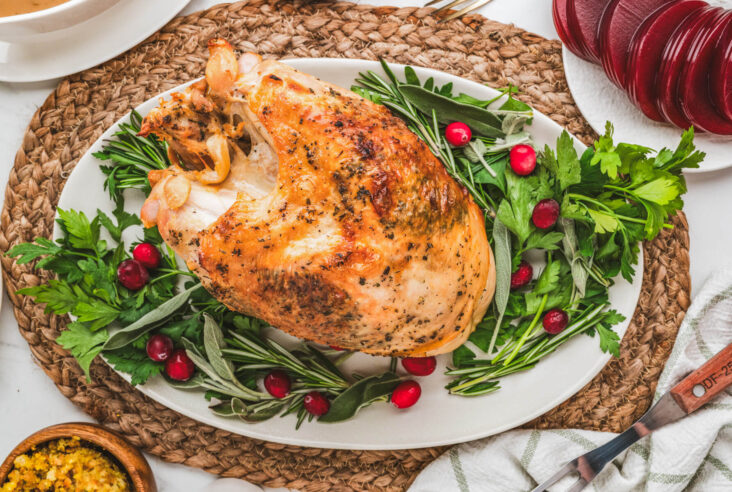 A golden brown crispy skin turkey breast on an oval platter surrounded by fresh herbs and cranberries.