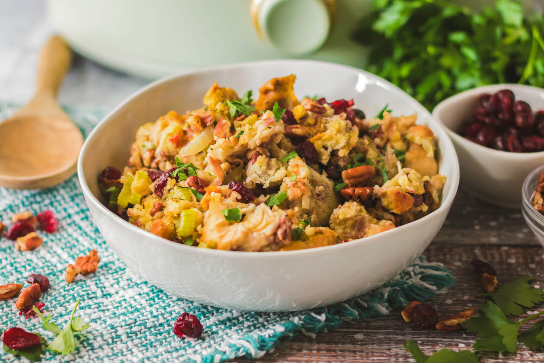 A white bowl with traditional bread dressing dotted with red dried cranberries, pecans, celery, and parsley in front of a slow cooker.