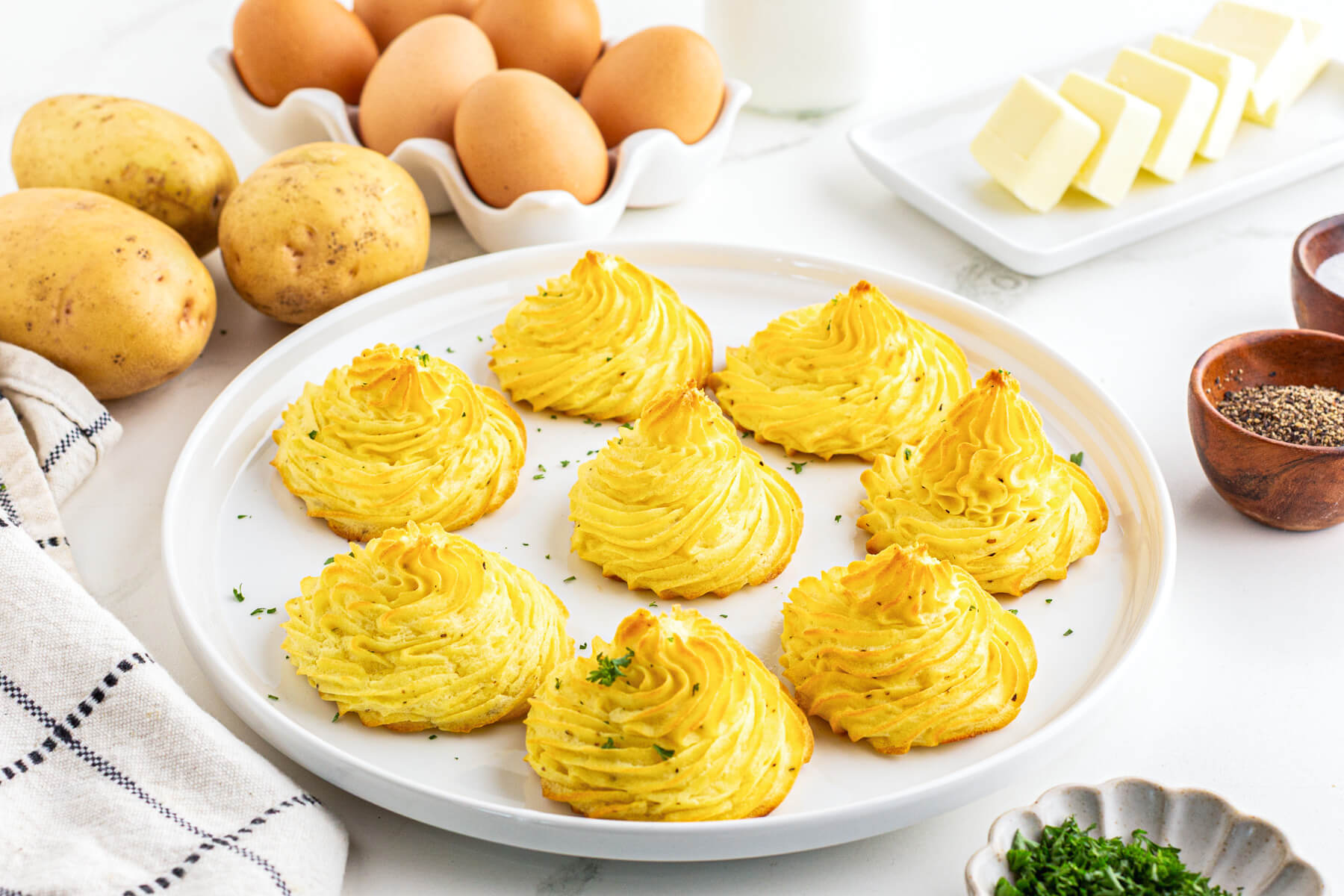 Golden baked swirls of rich baked Pommes Duchess on a white plate in front of a basket of brown eggs.