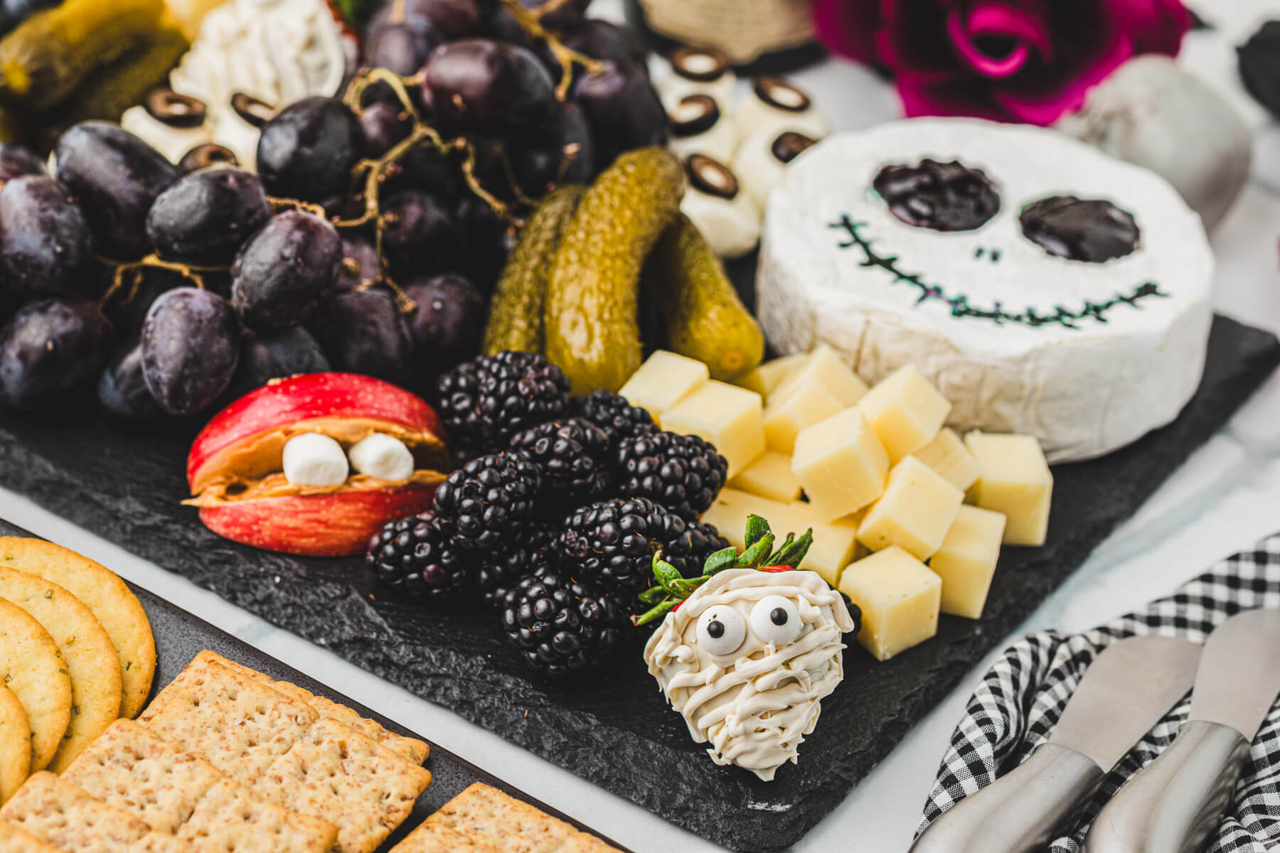 A Jack Skellington shaped brie cheese surrounded by pickles, black grapes, blackberries, apple mouths with fangs, cheese cubes, and other eery delights on this Halloween Charcuterie board.