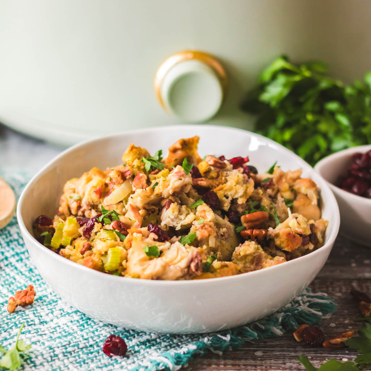 A white bowl with traditional bread dressing dotted with red dried cranberries, pecans, celery, and parsley in front of a slow cooker.