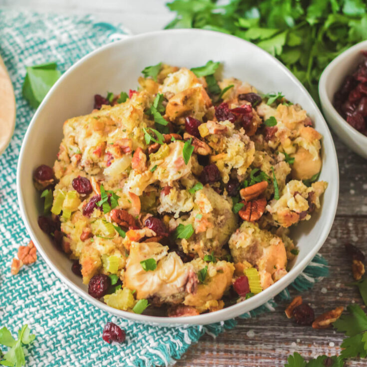 A white bowl with traditional bread dressing dotted with red dried cranberries, pecans, celery, and parsley.