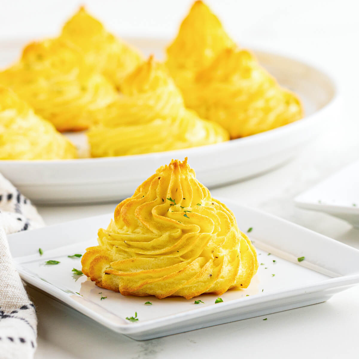 One Pomme Duchesse in front of white plate full of golden baked swirls of rich baked Duchess Potatoes.