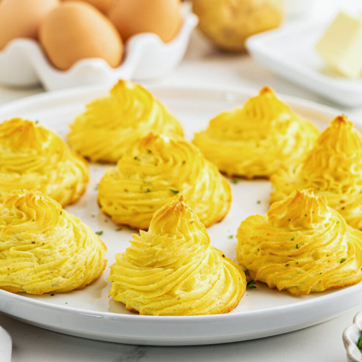 Golden baked swirls of rich baked Pommes Duchess on a white plate in front of a basket of brown eggs.
