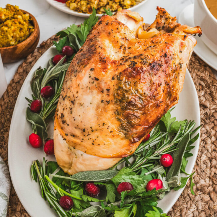 A golden brown crispy skin turkey breast on an oval platter surrounded by fresh herbs and cranberries.