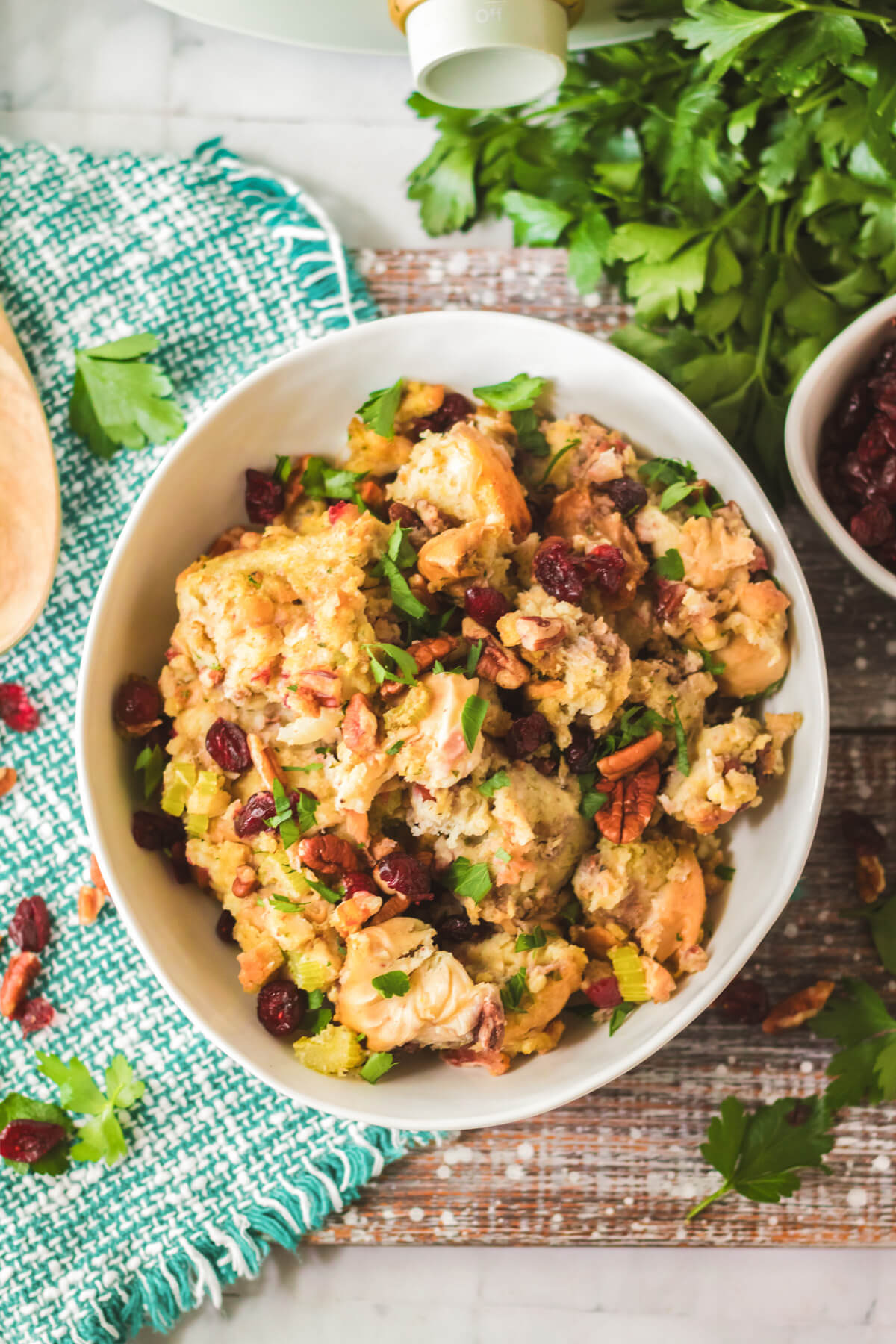 A white bowl with traditional bread dressing dotted with red dried cranberries, pecans, celery, and parsley.