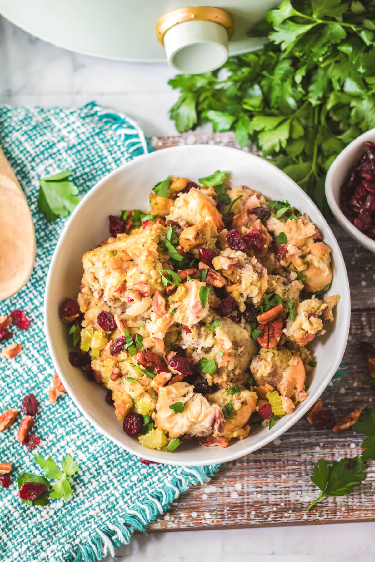 A white bowl with traditional bread dressing dotted with red dried cranberries, pecans, celery, and parsley in front of a slow cooker.