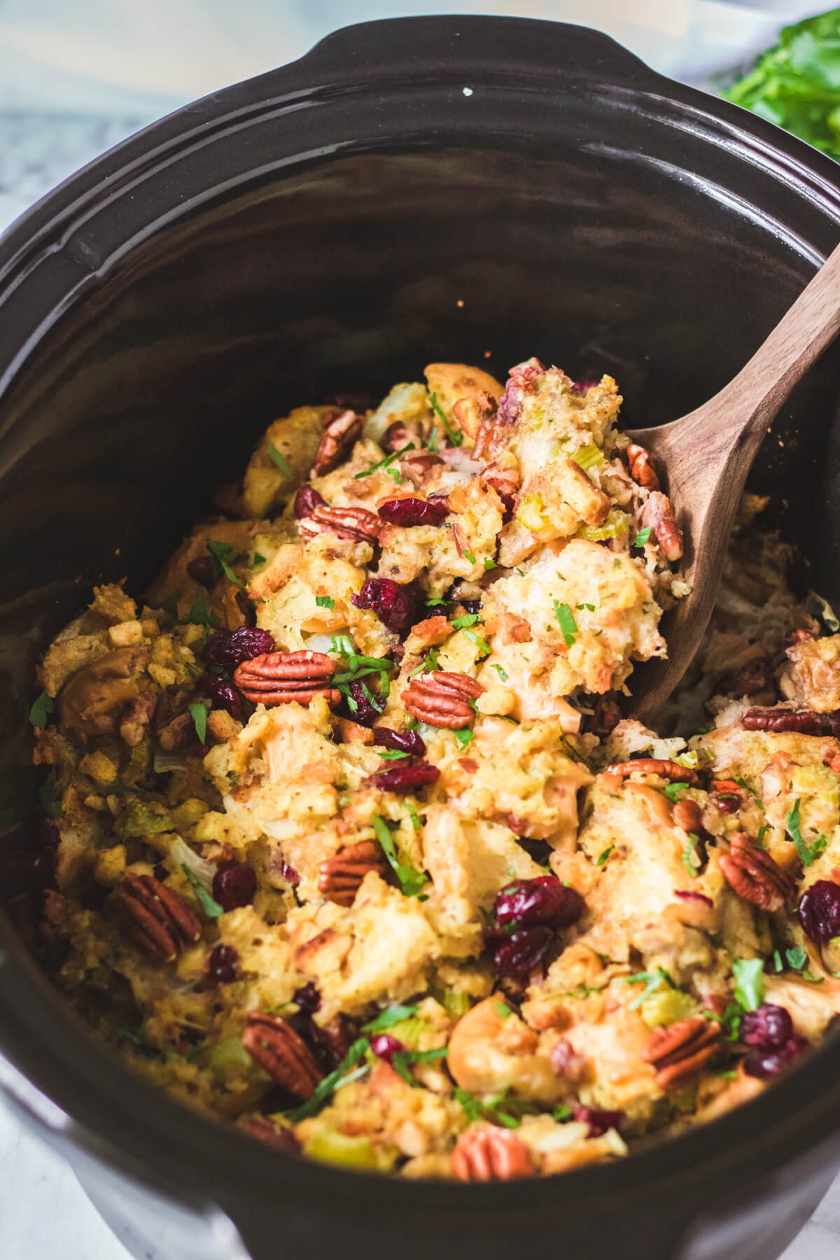 A wooden spoonful of Slow Cooker Dressing over a black slow cooker filled with traditional bread dressing dotted with red dried cranberries, pecans, celery, and parsley.