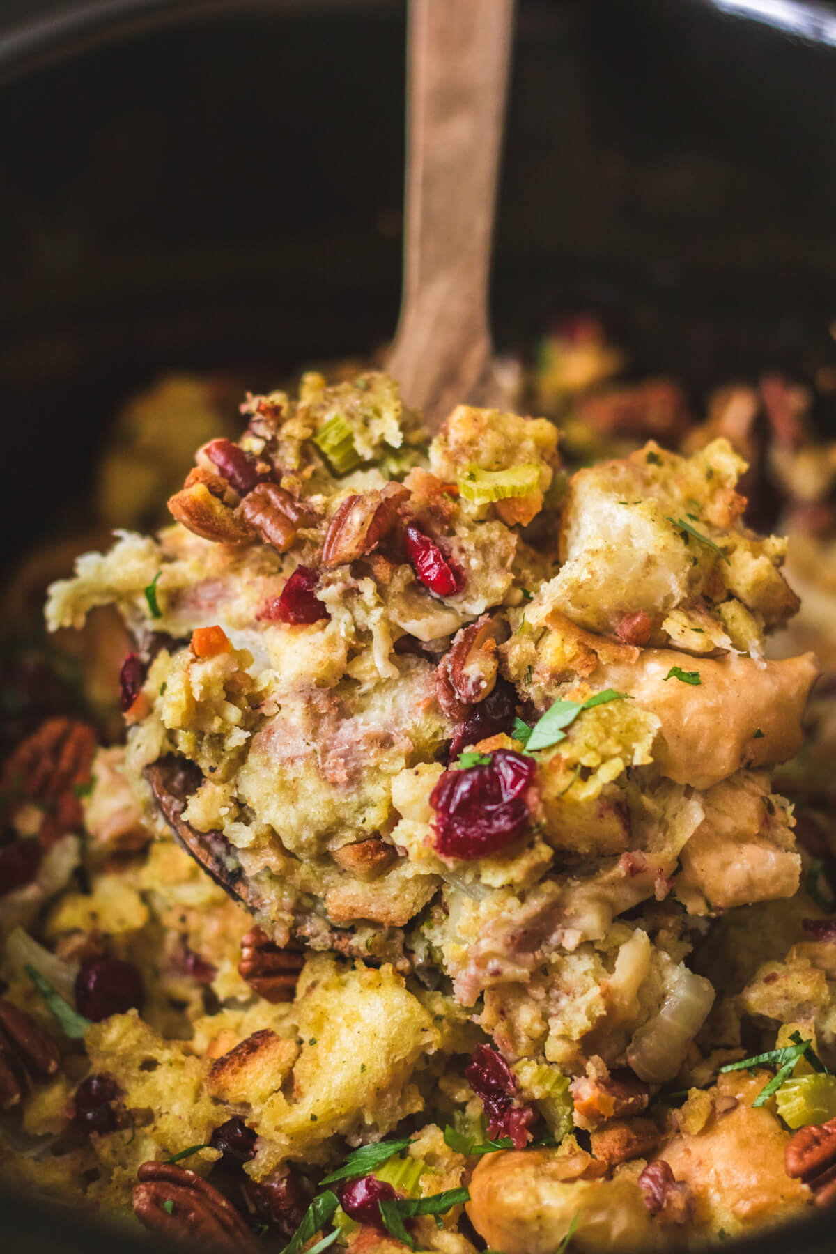 A wooden spoonful of Slow Cooker Dressing over a black slow cooker filled with traditional bread dressing dotted with red dried cranberries, pecans, celery, and parsley.