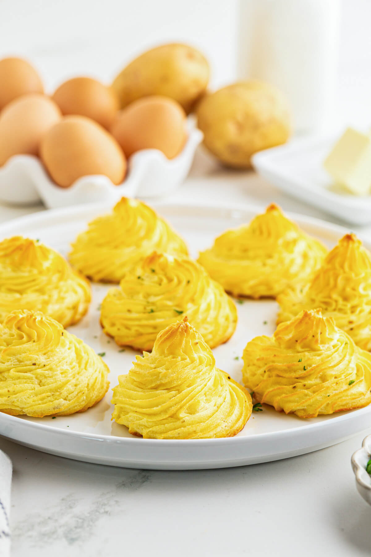 Golden baked swirls of rich baked Pommes Duchess on a white plate in front of a basket of brown eggs.