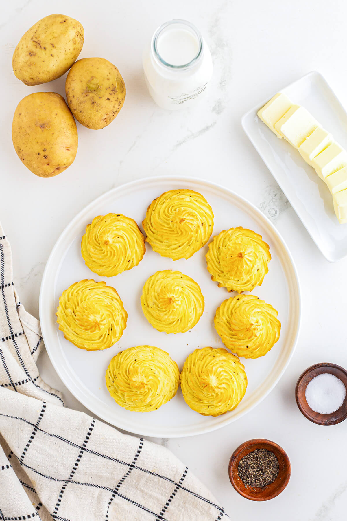 Golden baked swirls of rich baked Pommes Duchess on a white plate surrounded by recipe ingredients.