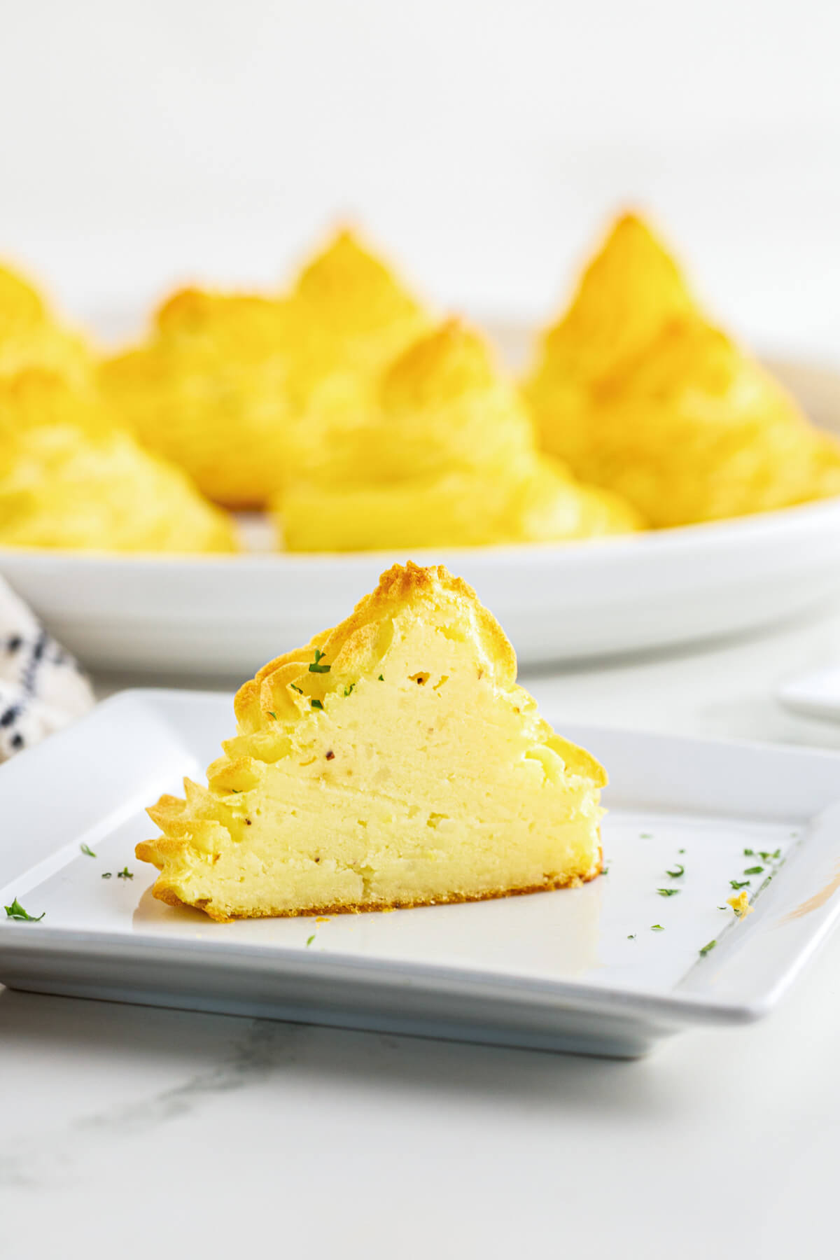 One sliced Pomme Duchesse in front of white plate full of golden baked swirls of rich baked Duchess Potatoes.