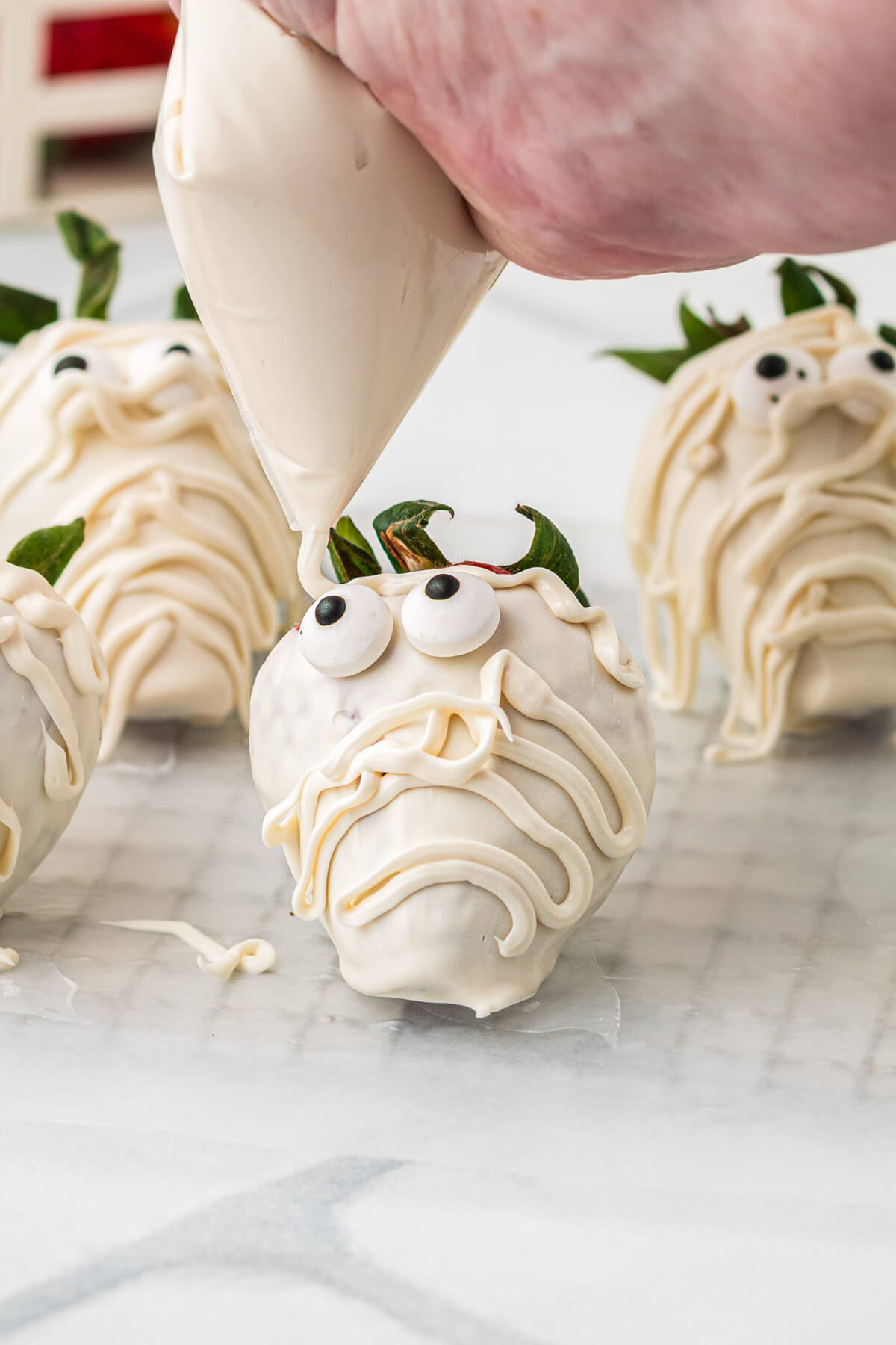 A hand drizzles melted white chocolate onto a white chocolate strawberry with google eyes to make it into a mummy.