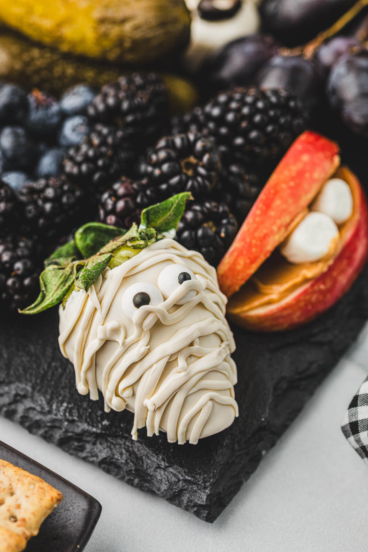 A fanged mouth made of apples and mini marshmallows and mummified strawberry beside blackberries and black grapes on a Halloween Charcuterie Board.