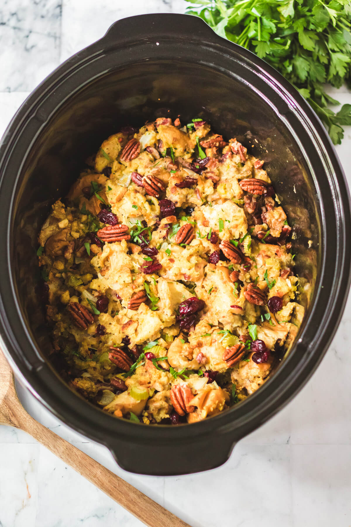Slow Cooker Dressing in a black slow cooker with traditional bread dressing dotted with red dried cranberries, pecans, celery, and parsley.