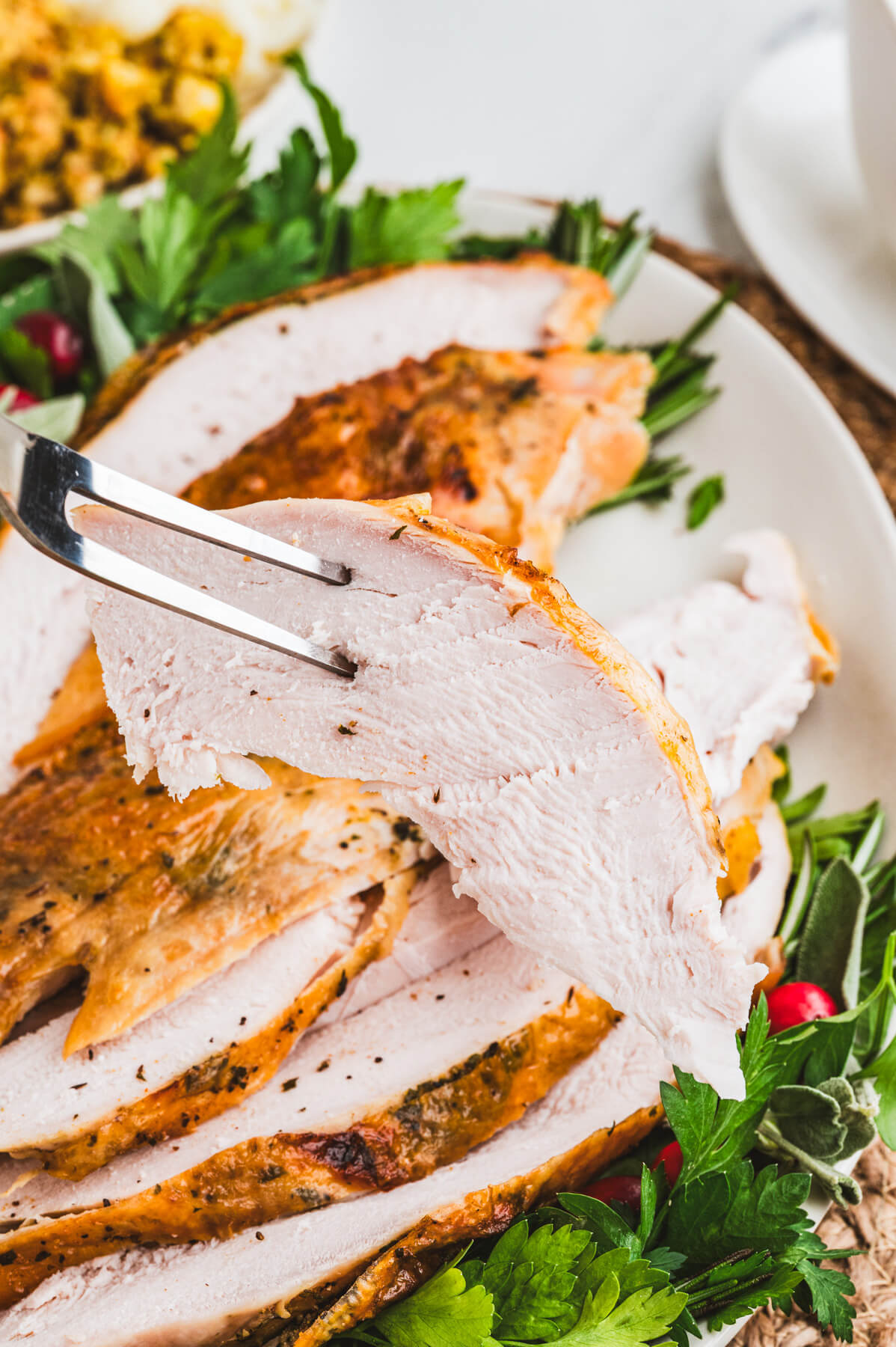 A carving fork holds a slice of turkey breast. above a dinner plate.