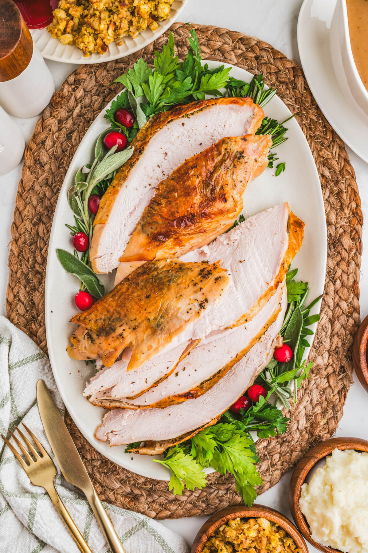 A sliced golden brown crispy skin turkey breast on an oval platter surrounded by fresh herbs and cranberries.