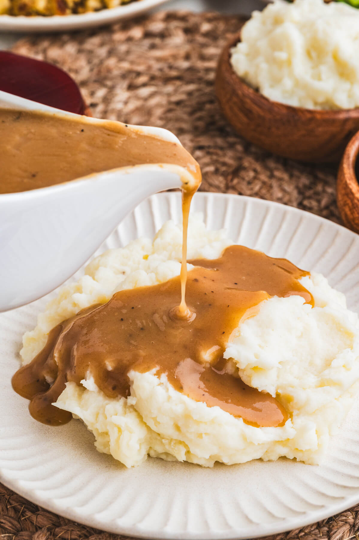 Rich brown gravy pours from a white gravy boat over a plate of mashed potatoes.
