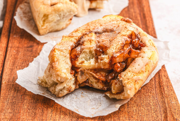 Two golden baked Apple Pie Cinnamon Rolls drizzled with frosting on a wooden serving board.