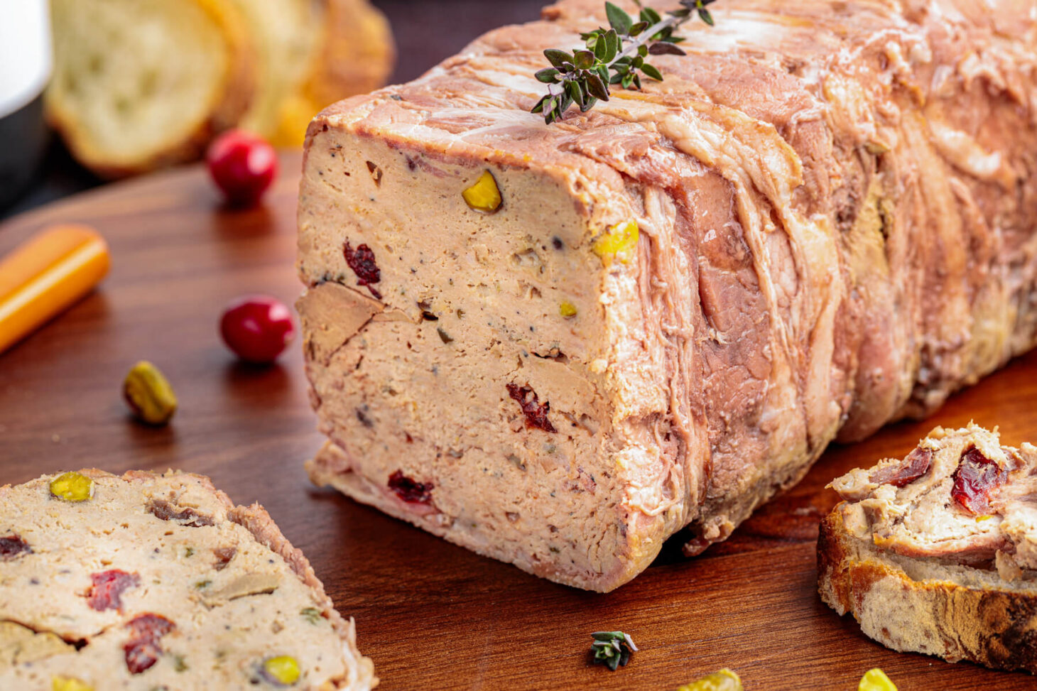 A sliced bacon wrapped pork terrine dotted with dried cranberries and green pistachios on a cutting board.