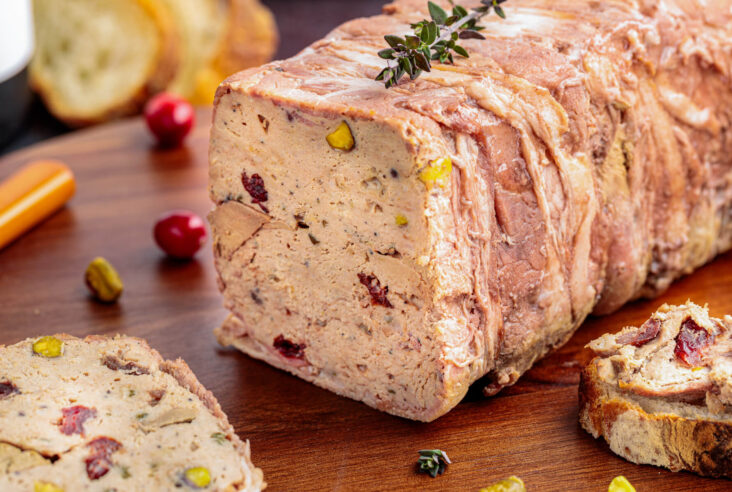 A sliced bacon wrapped pork terrine dotted with dried cranberries and green pistachios on a cutting board.