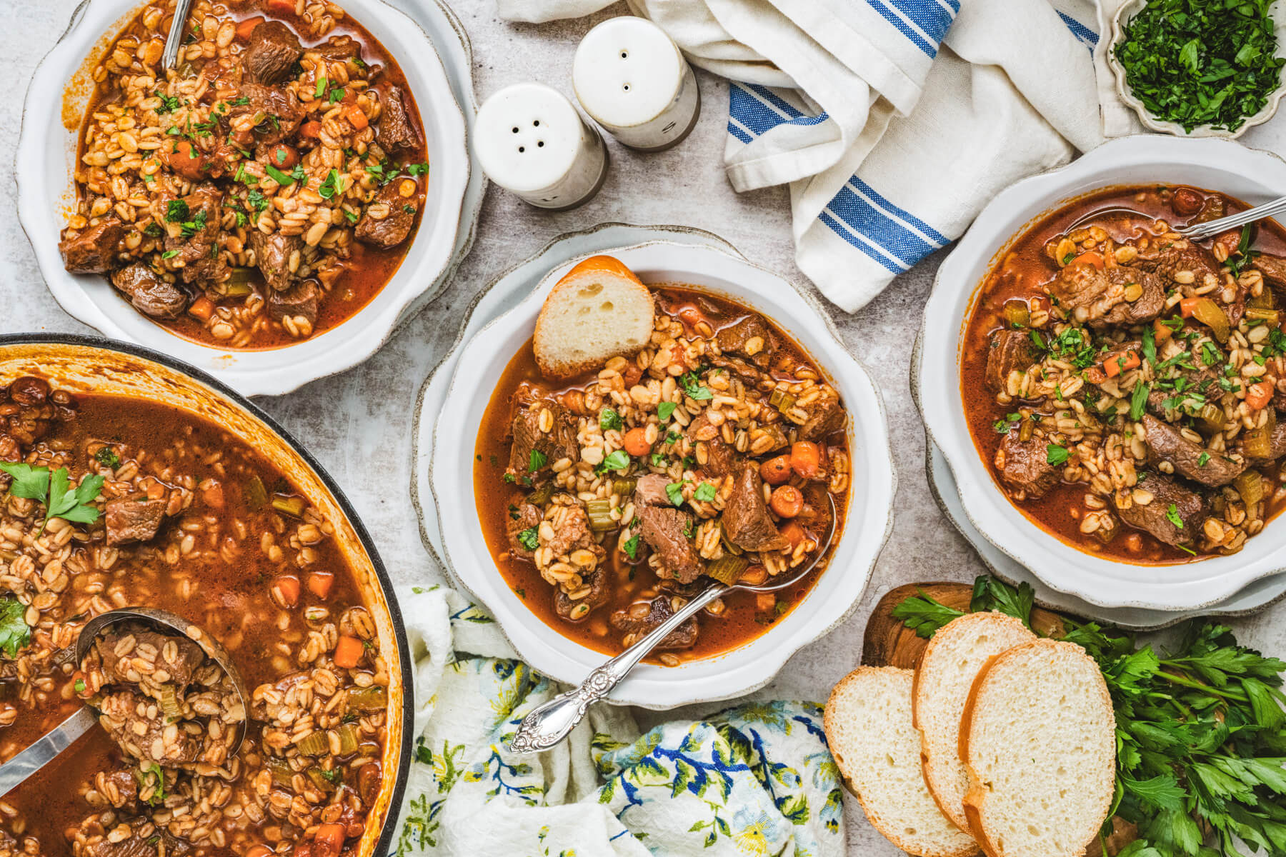 A table scene featuring white bowls and a large pot filled with beef barley soup beside slices of bread and fresh herbs.