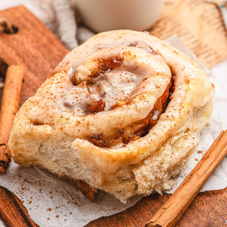 One golden baked glazed Apple Pie Cinnamon Roll on a wooden board surrounded by cinnamon sticks.