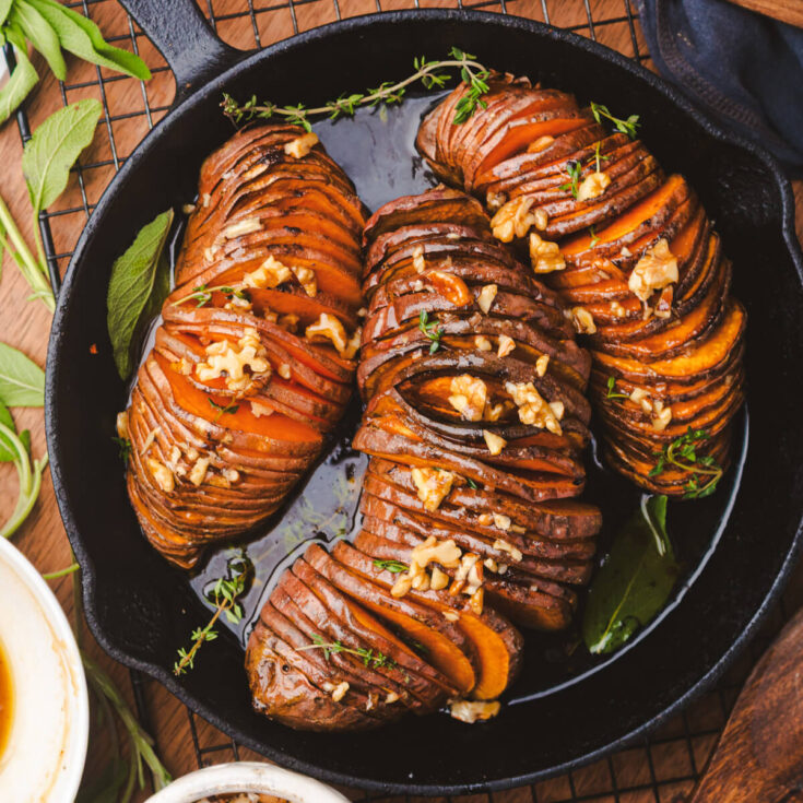 Three perfectly roasted vibrant orange hasselback sweet potatoes topped with herbs and walnuts in a cast iron pan.