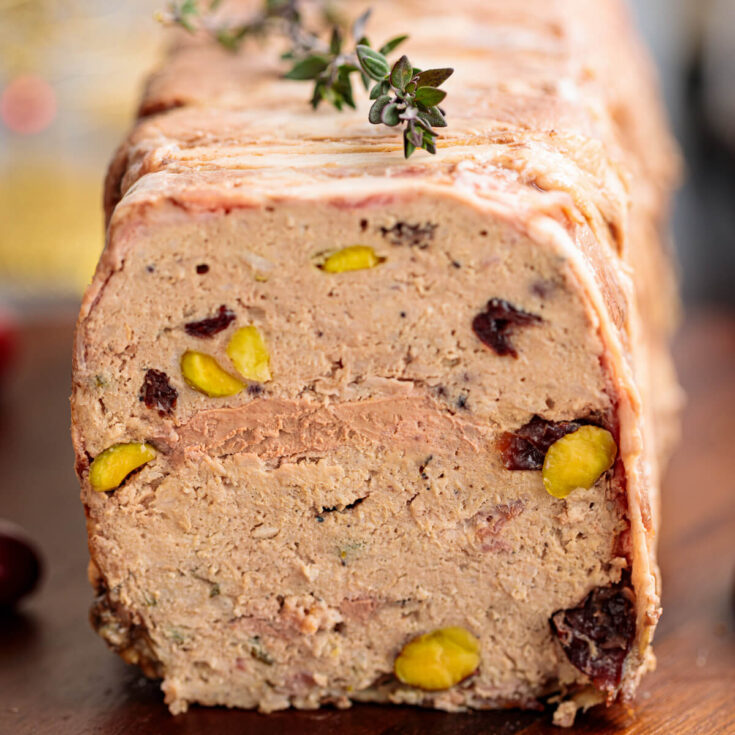 A sliced bacon wrapped pork terrine dotted with dried cranberries and green pistachios on a cutting board.