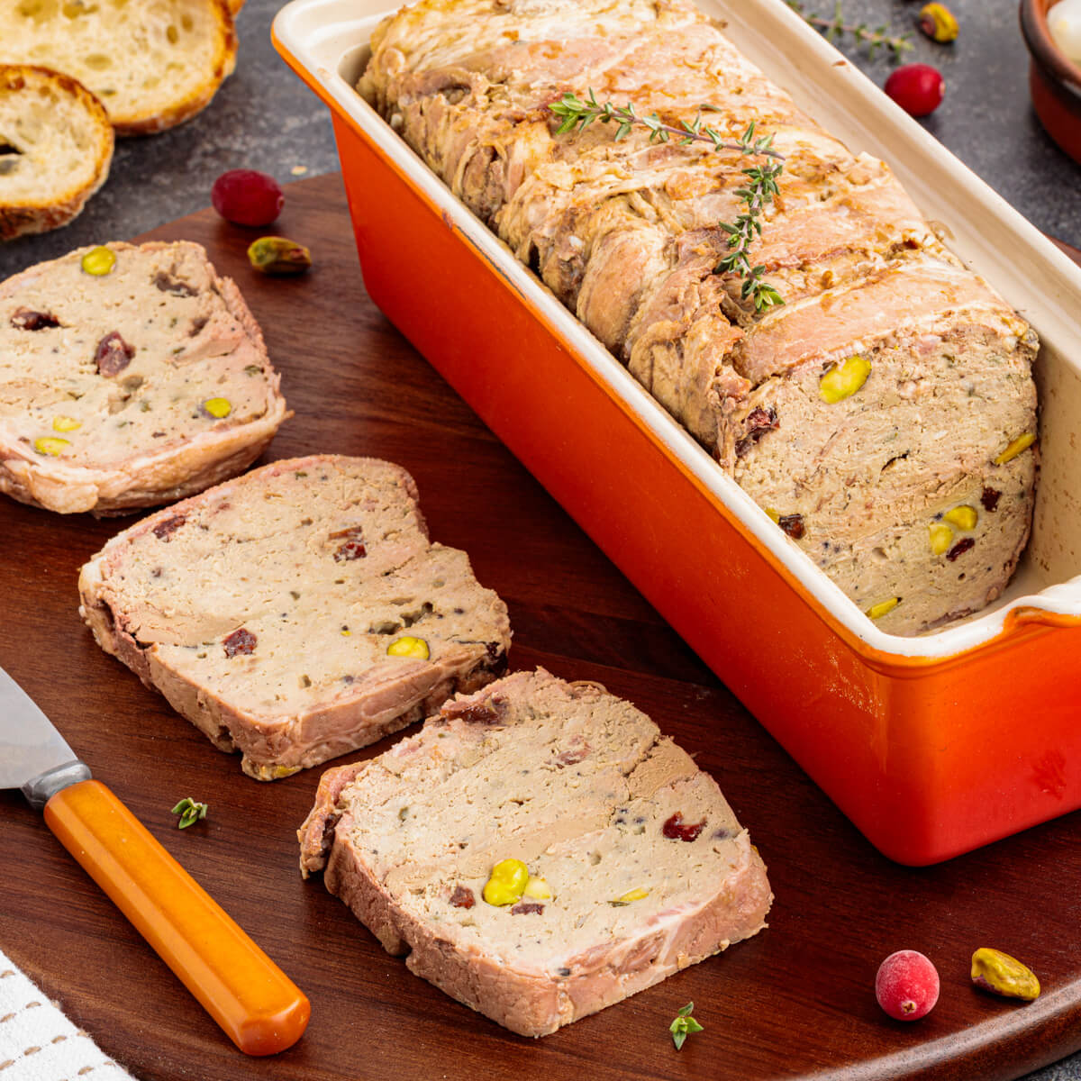 Three slices of bacon wrapped pork terrine dotted with dried cranberries and green pistachios on a cutting board beside a red terrine pan.