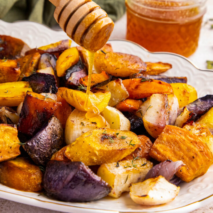 Colourful roasted root vegetables on a white platter being drizzled with honey