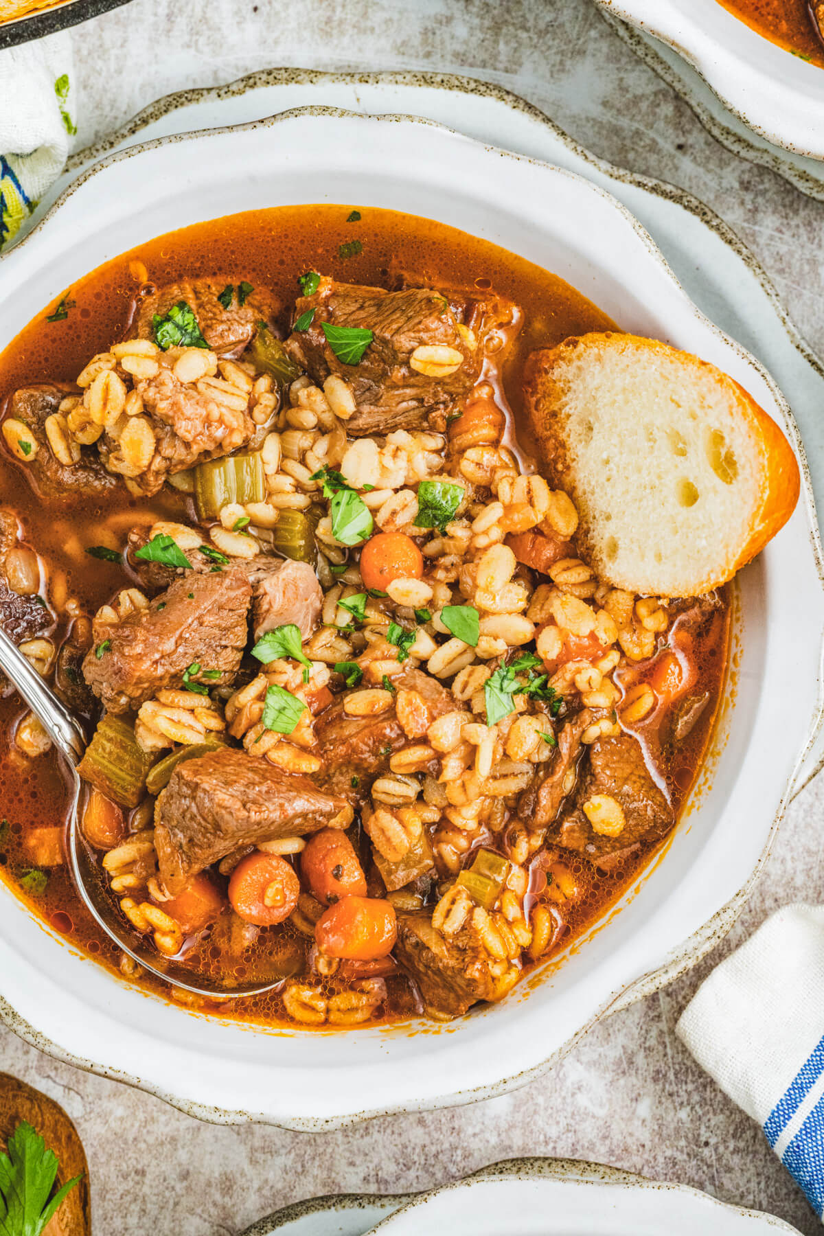 A white bowl filled with beef barley stew garnished with fresh parsley and a slice of bread.