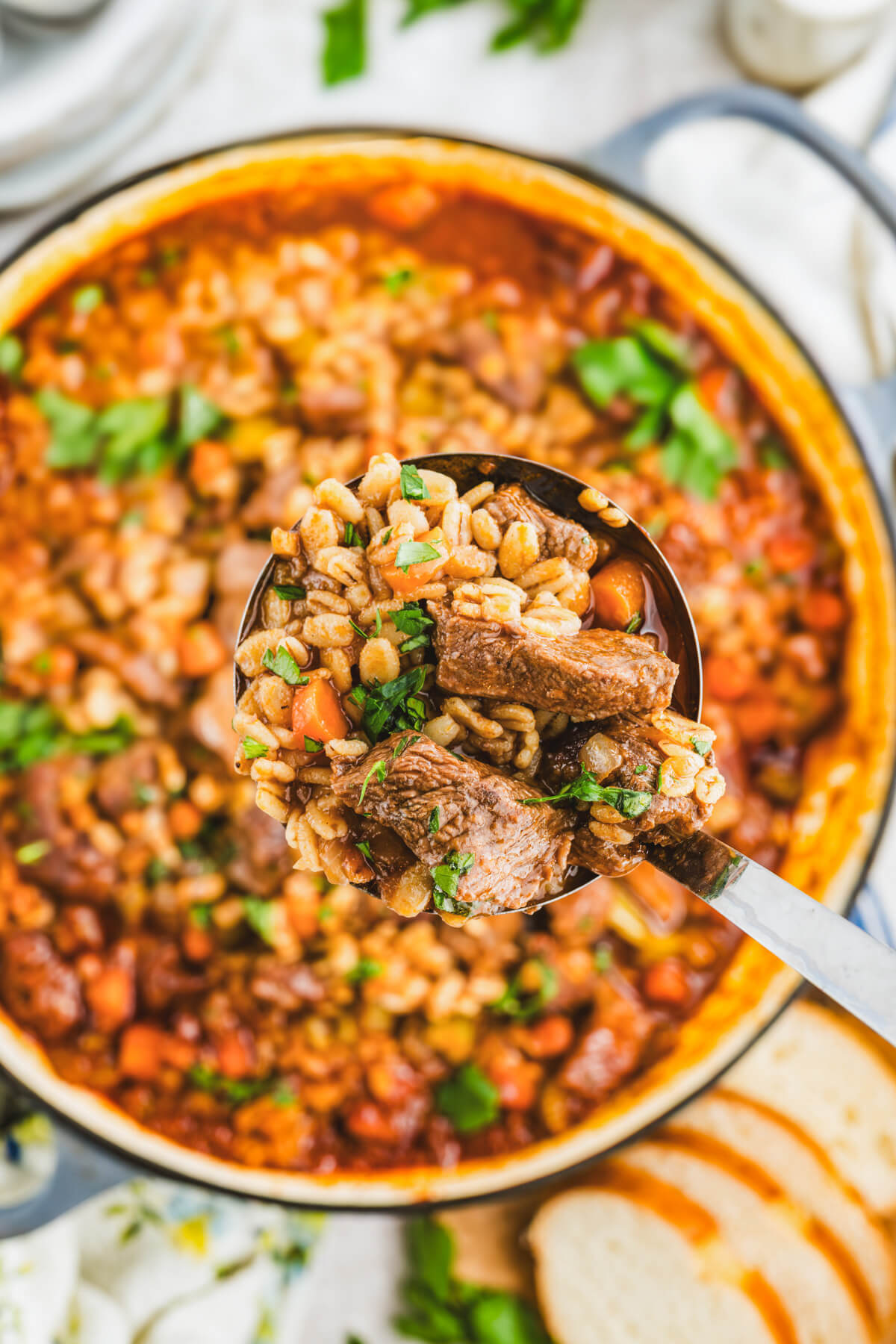 A serving spoon holding chunky beef barley stew over a large pot filled with the same stew.