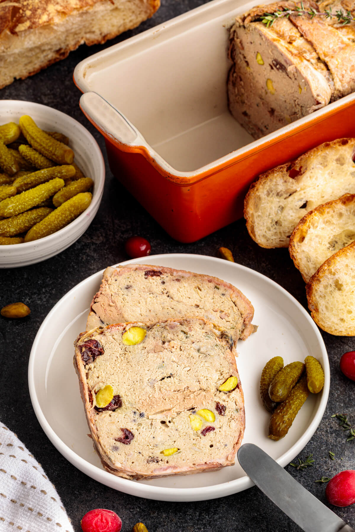 Slices of bacon wrapped pork terrine dotted with dried cranberries and green pistachios on white plate.