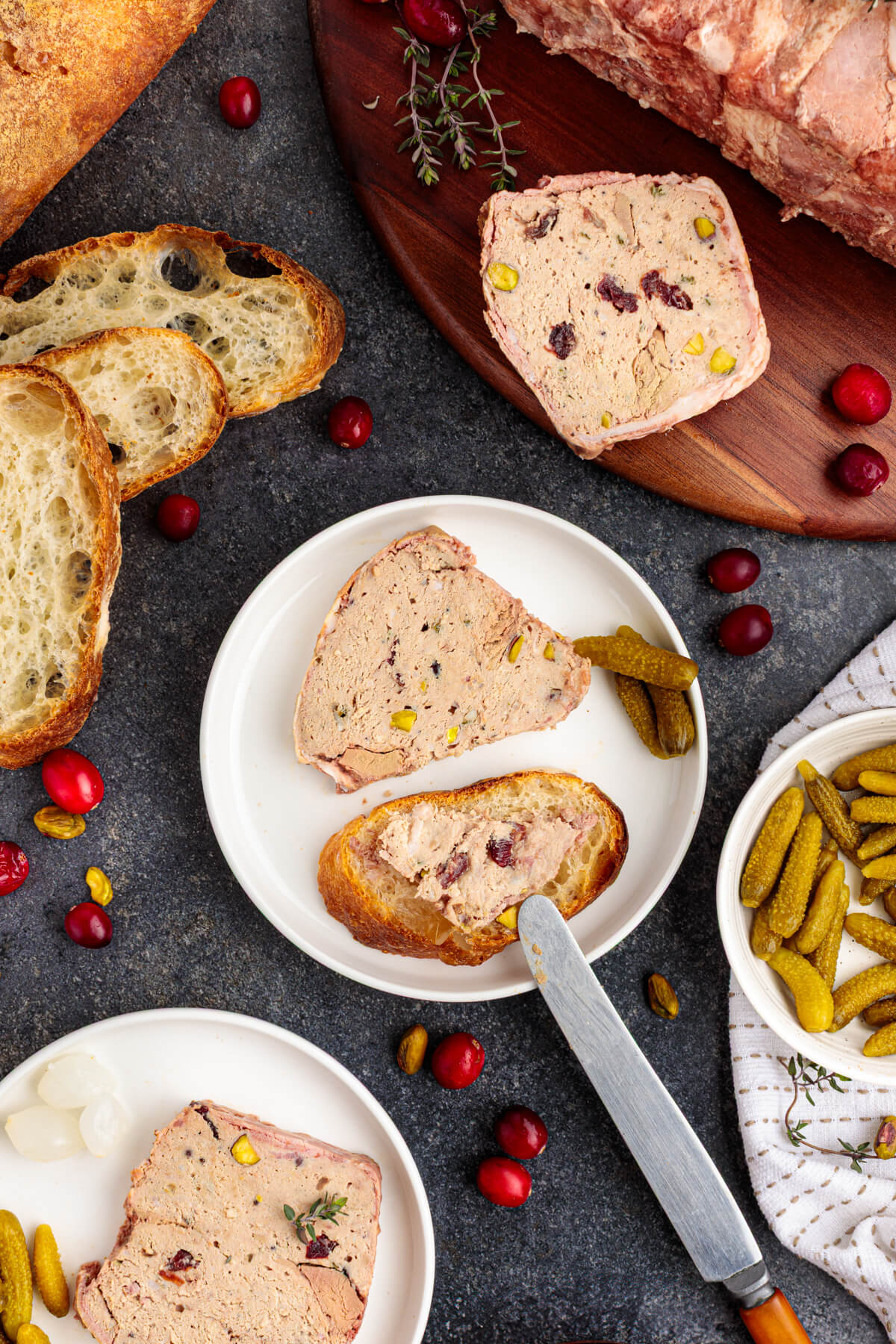 A knife spreads pork terrine dotted with dried cranberries and green pistachios on a slice of baguette.