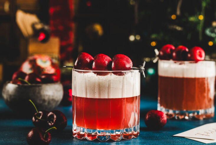 Two rocks glasses filled with vibrant red amaretto cherry sour cocktail with top layers of foam and three cherries as garnish.
