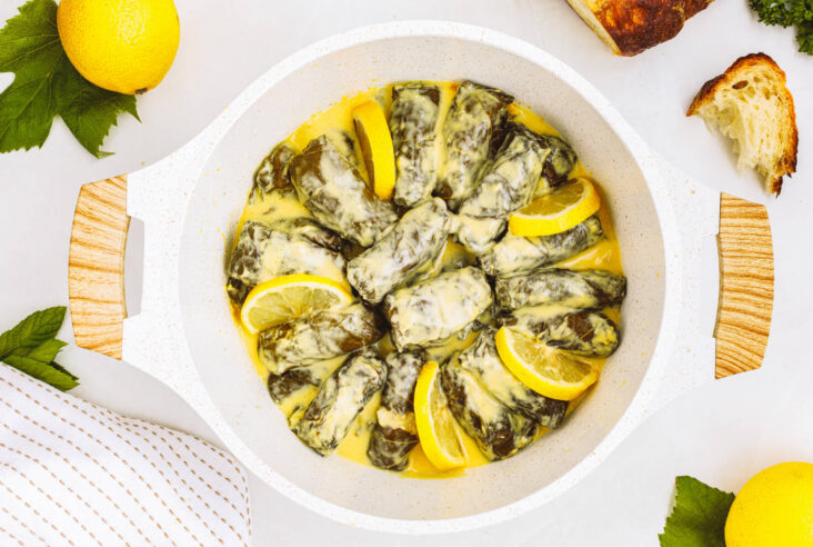 A round roasting pan full of stuffed grape vine leaves with lemon slices in a creamy yellow sauce.