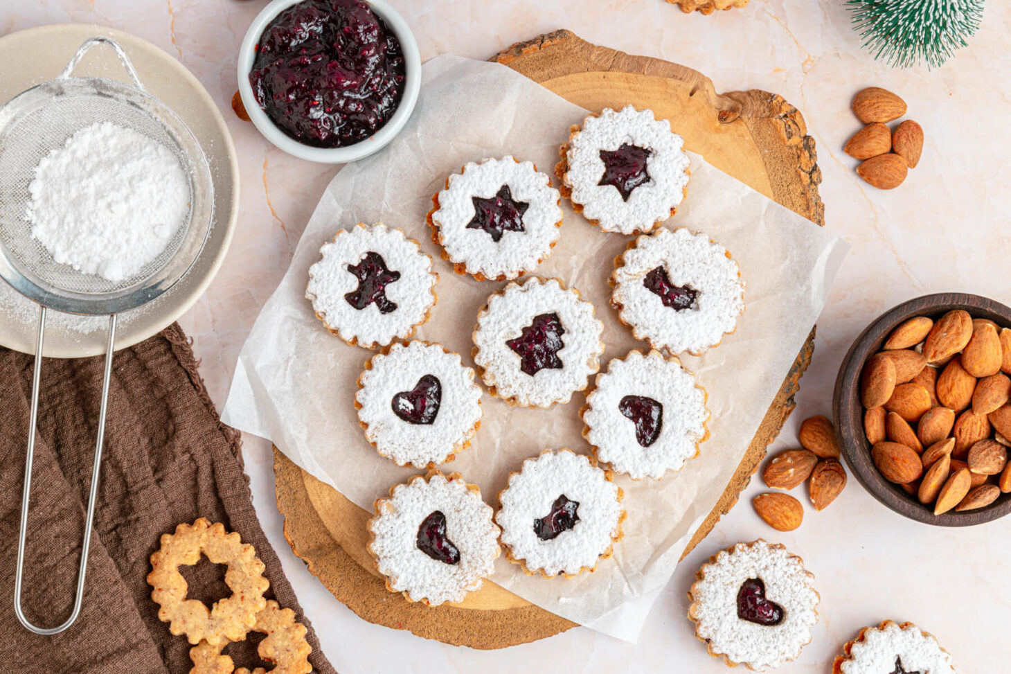 Raspberry Linzer sandwich cookies with bell, star, and heart shaped cut outs dusted in confectioner's sugar on a wooden board.