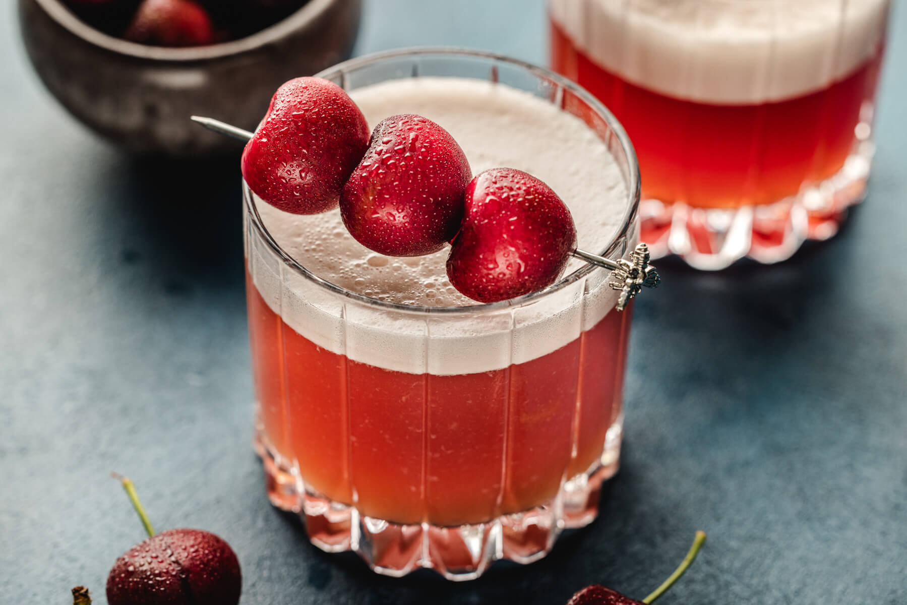 Two rocks glasses filled with vibrant red amaretto cherry sour cocktail with top layers of foam and three cherries as garnish.