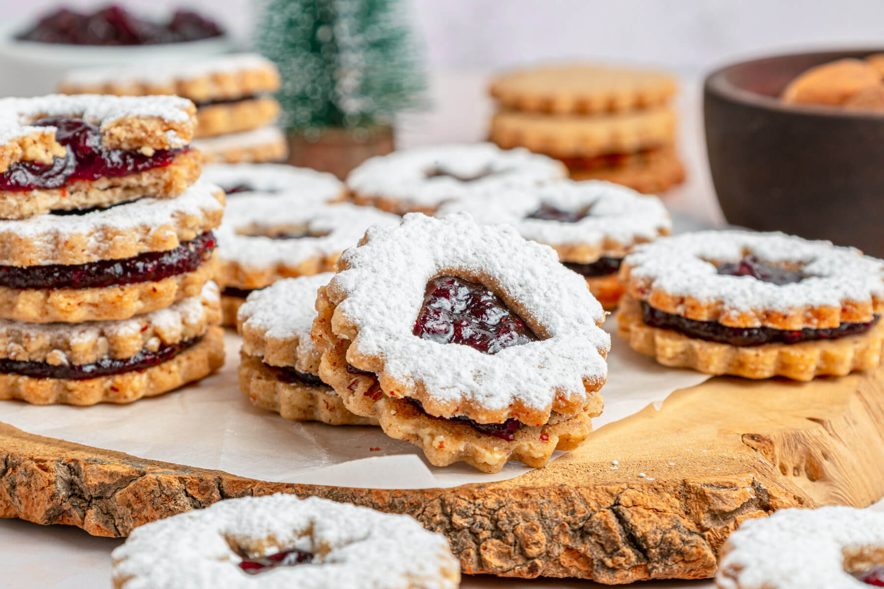 Raspberry Linzer sandwich cookies with bell, star, and heart shaped cut outs dusted in confectioner's sugar on a wooden board.