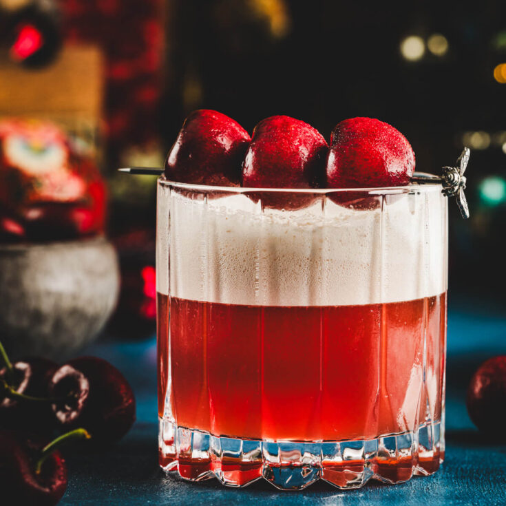 A rocks glass filled with vibrant red amaretto cherry sour cocktail with a top layer of foam and three cherries as garnish.