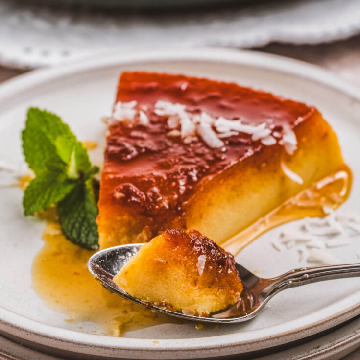 A spoon contains a bite of flan beside a slice of creamy coconut flan topped with a golden brown caramel and coconut flakes.