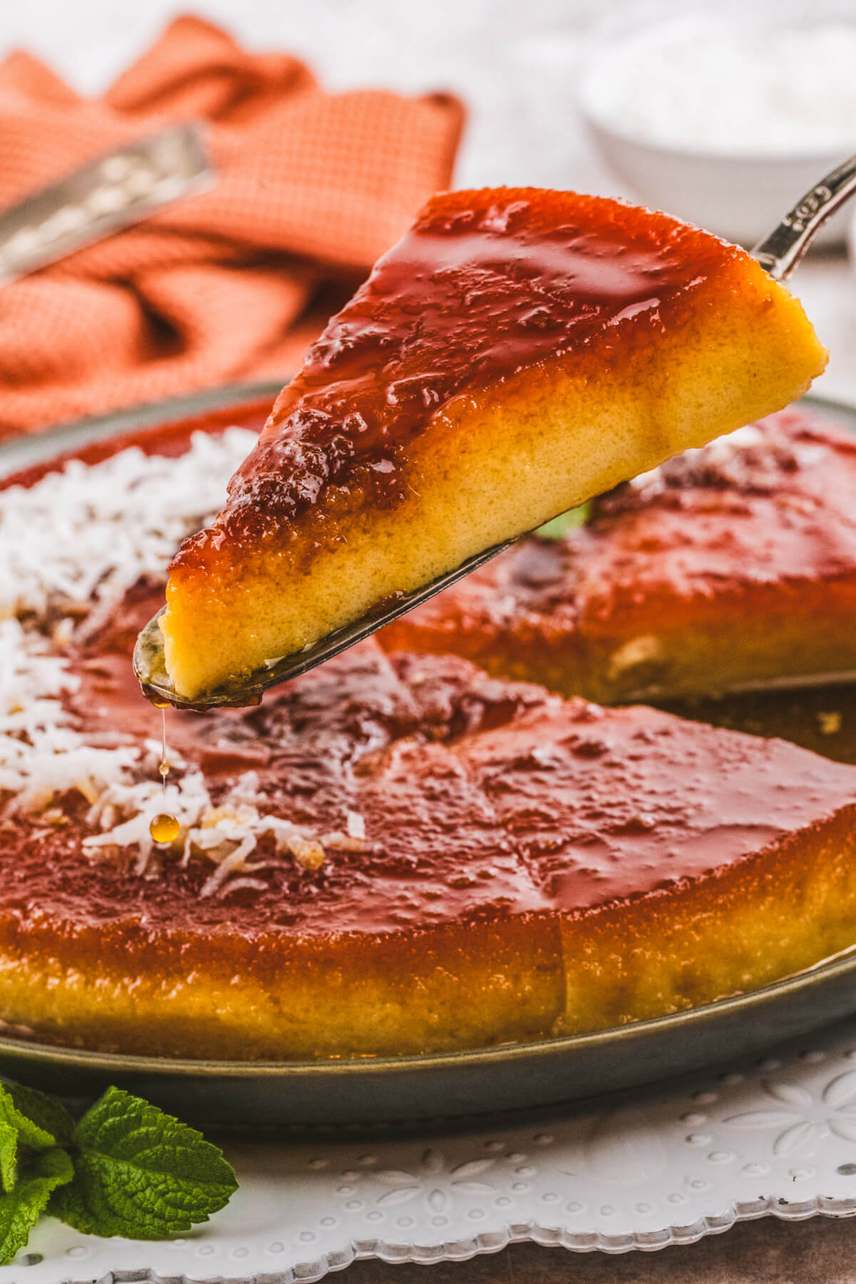 A cakes server holds a slice of coconut flan above a whole flan topped with a golden brown caramel and coconut flakes.