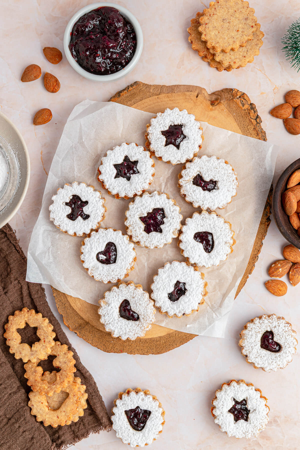 Raspberry Linzer sandwich cookies with bell, star, and heart shaped cut outs dusted in confectioner's sugar on a wooden board.