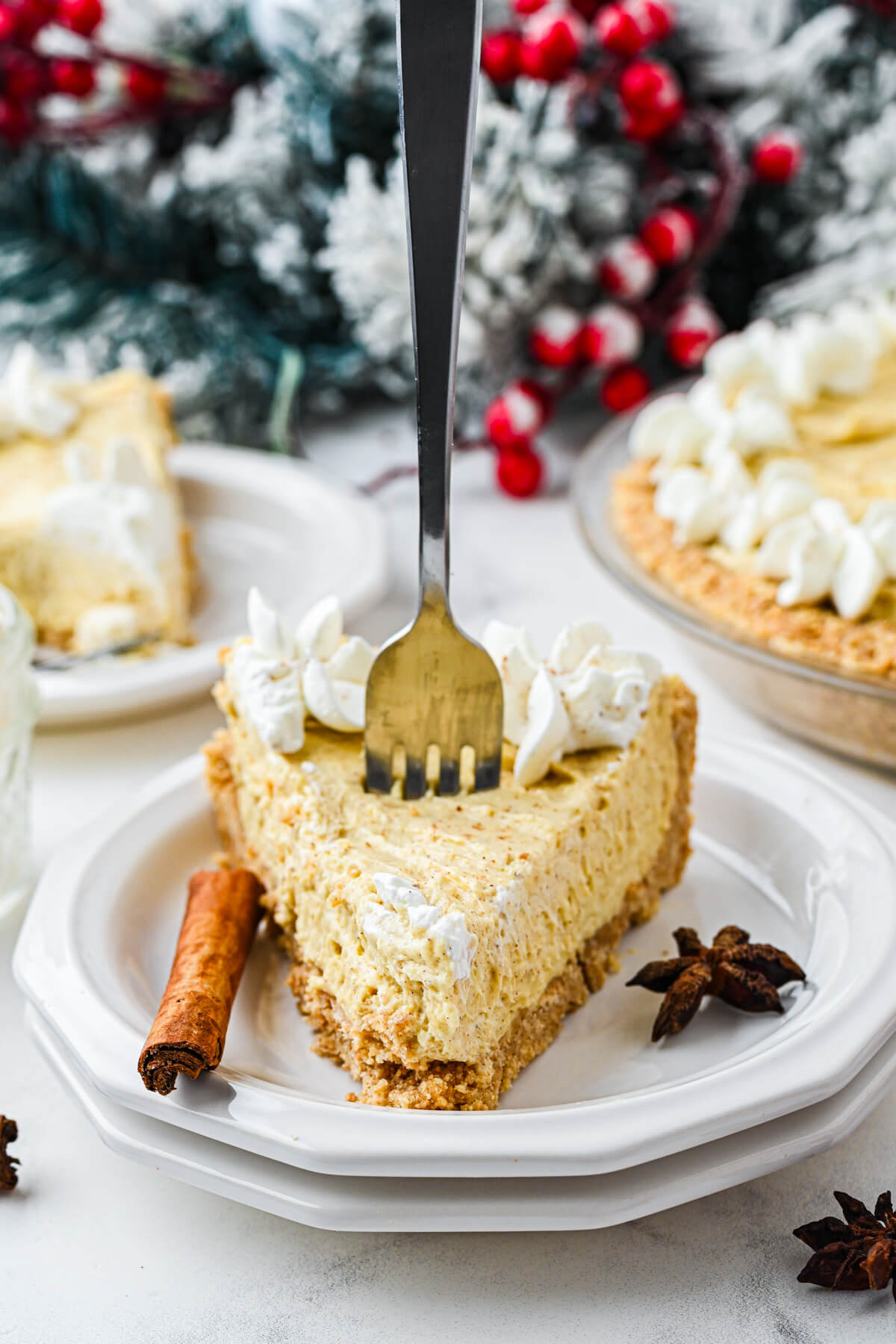 A fork stands upright in a slice of No Bake Eggnog Pie on a white plate with a cinnamon stick and star anise to decorate.