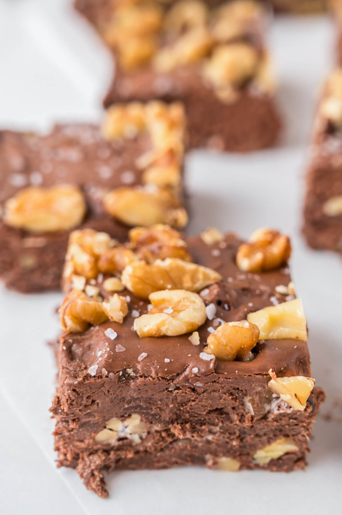 A group of chocolate walnut fudge squares topped with chopped walnuts and sea salt on a white plate.